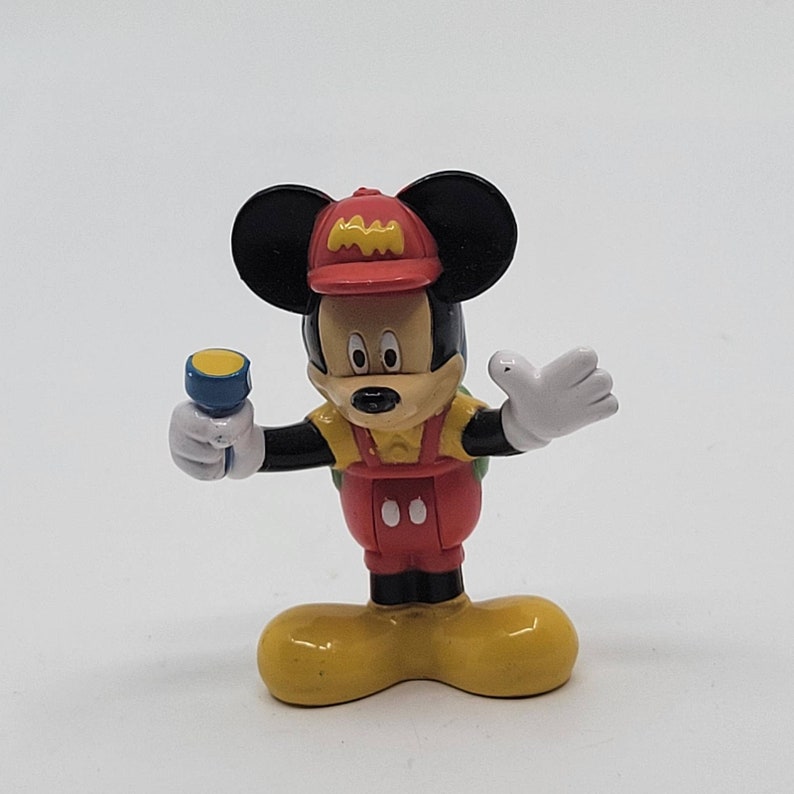 May include: A red, yellow and black Mickey Mouse figurine wearing a red hat with the letters "MM" on it. The figurine is holding a blue torch and has a green backpack.