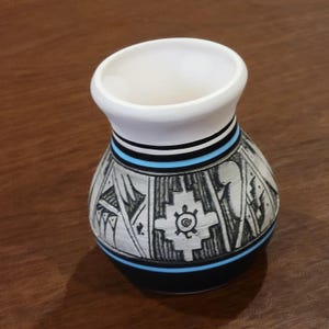 May include: A handcrafted ceramic vase with a white interior and a black and white exterior. The vase features a detailed geometric design with a turquoise and black band around the neck. The base is black.
