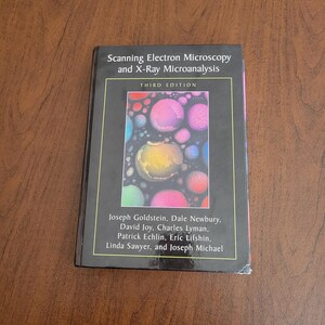 Peut inclure: Un livre à couverture rigide intitulé "Scanning Electron Microscopy and X-Ray Microanalysis, Third Edition". La couverture présente un cadre noir et un motif abstrait coloré de formes circulaires. Les noms des auteurs sont listés en dessous.