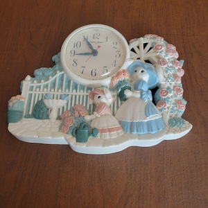 May include: A decorative wall clock depicting a garden scene with two figures, a white picket fence, and a floral arch. The clock face is white with black numerals and hands. The figures are dressed in pastel colours, and the design is whimsical.