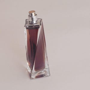 Puede incluir: Un frasco de perfume rectangular con tapón plateado. El frasco es transparente con un líquido burdeos oscuro en su interior. El diseño presenta facetas geométricas anguladas, creando un aspecto moderno y elegante. El frasco está sobre una superficie blanca.