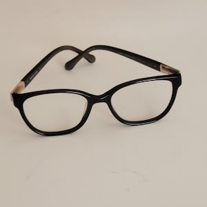 May include: Black eyeglasses with a cat-eye frame design. The glasses have a glossy finish and feature a thin, dark frame with a subtle gold accent near the hinges. The lenses are clear, and the temples curve gently.