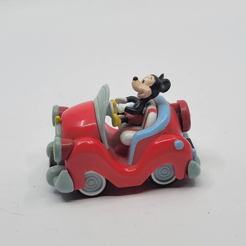 Puede incluir: Un coche de juguete rojo con una figura de Mickey Mouse al volante. El coche tiene detalles azules y blancos y ruedas negras.