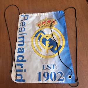 Könnte beinhalten: Weiß-blauer Turnbeutel mit dem Real Madrid-Logo. Das Logo ist ein goldenes Emblem mit Krone und den Buchstaben "MCF". Der Text "Realmadrid" ist vertikal links und "EST. 1902" unten aufgedruckt.
