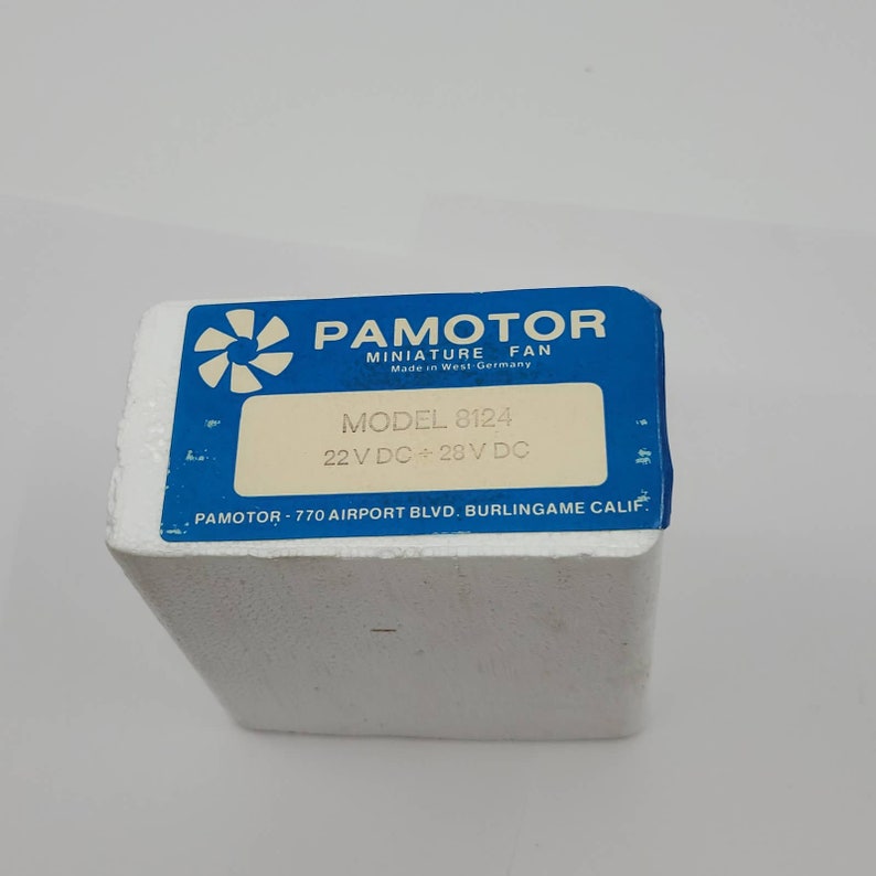 May include: A blue and white box labeled "PAMOTOR MINIATURE FAN" with the text "Made in West Germany." The box has a label that reads "MODEL 8124" and "22 V DC + 28 V DC." The box is on a white surface.
