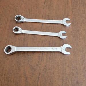 Puede incluir: Tres llaves de carraca reversibles plateadas con extremos abiertos, dispuestas sobre una superficie marrón. Las llaves tienen diferentes tamaños, incluyendo 5/8, 1/2 y 7/16 pulgadas. La marca "Doverable GoldWrench" es visible en una llave.