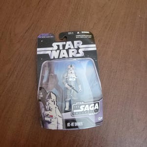 May include: A Star Wars Saga Collection action figure of an AT-AT Driver in a white and gray uniform with a blaster rifle. The figure is packaged in a plastic blister pack with a blue background and the Star Wars logo.