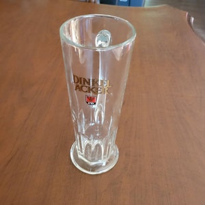 May include: A clear glass beer stein with a gold logo that reads "Dinkel Acker" and a red and gold crest. The stein has a handle and a textured base.