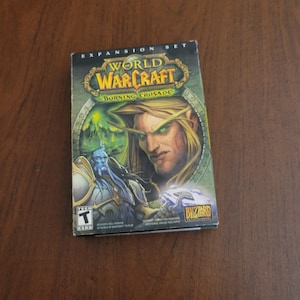 Może przedstawiać: Pudełko z grą rozszerzającą World of Warcraft: Burning Crusade. Pudełko ma zielono-złoty design z ilustracjami postaci. Logo Blizzard jest widoczne w prawym dolnym rogu. Gra ma ocenę T dla nastolatków.