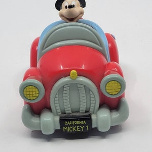 Puede incluir: Un coche de juguete rojo con una figura de Mickey Mouse al volante. El coche tiene un parabrisas azul, faros amarillos y una matr&iacute;cula que dice "CALIFORNIA MICKEY 1".