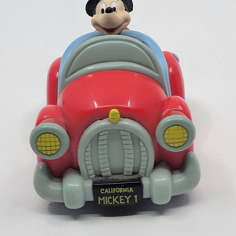 Puede incluir: Un coche de juguete rojo con una figura de Mickey Mouse al volante. El coche tiene un parabrisas azul, faros amarillos y una matr&iacute;cula que dice "CALIFORNIA MICKEY 1".