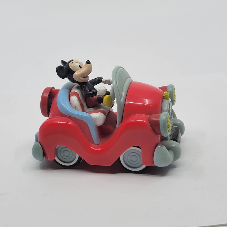 Puede incluir: Un coche de juguete rojo con una figura de Mickey Mouse al volante. El coche tiene detalles azules y blancos y faros amarillos.