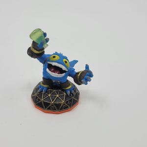 May include: A blue action figure of a cartoonish creature with yellow eyes, a wide open mouth, and a thumbs-up gesture. The figure is holding a green translucent object and stands on a black and orange base.