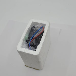 May include: A black rectangular object wrapped in clear plastic, nestled inside a white foam container. Red and blue wires extend from the object. The container is rectangular with a slightly angled top.