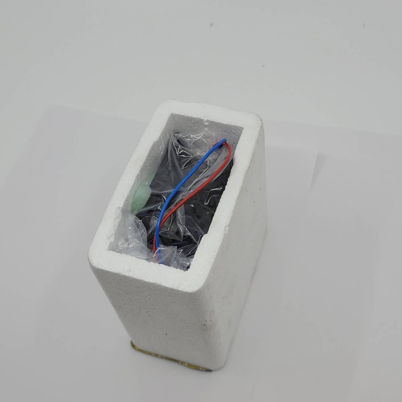May include: A black rectangular object wrapped in clear plastic, nestled inside a white foam container. Red and blue wires extend from the object. The container is rectangular with a slightly angled top.