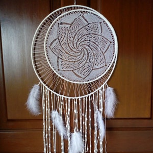 Puede incluir: Un atrapasueños blanco con un diseño circular, con un patrón de telaraña detallado y de encaje dentro del círculo más grande. El atrapasueños tiene una forma de media luna debajo del círculo, con plumas blancas y hebras de cuentas colgando. El fondo es una puerta de madera marrón.