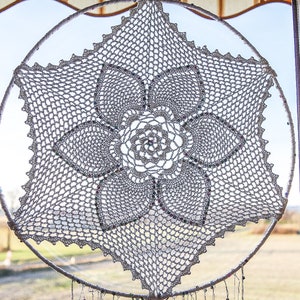 Puede incluir: Un atrapasueños de crochet blanco con flecos de cuentas colgando de una ventana. El atrapasueños tiene un gran diseño de flor intrincado en el centro.