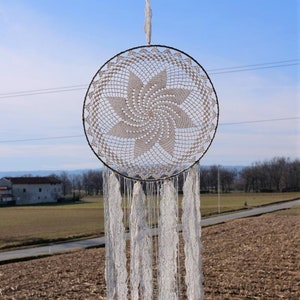 Puede incluir: Un atrapasueños de crochet blanco con un aro circular y largas flecos blancos colgando de una valla de madera.