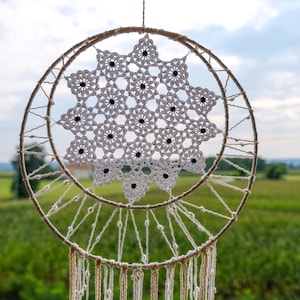 Puede incluir: Un atrapasueños de crochet blanco con cuentas negras colgando sobre un fondo de campo verde y cielo nublado.