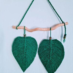 Puede incluir: Dos macramés de pared en forma de hoja verde con listones de madera y cuerda verde, colgando de una cuerda verde en una pared azul claro.