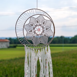 Puede incluir: Un atrapasueños de crochet blanco con cuentas rojas que cuelgan de un aro marrón. El atrapasueños está colgado de un toldo a rayas sobre un fondo de cielo azul y campo verde.