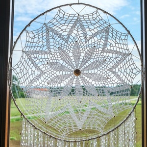 Puede incluir: Atrapasueños de crochet blanco con flecos de cuentas colgando en una ventana. El atrapasueños es circular y tiene un diseño intrincado.