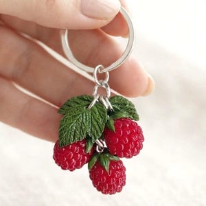 May include: A silver-toned metal keyring featuring three realistic, textured red raspberry charms attached to a circular split ring. The raspberries are clustered together with detailed green leaves, creating a lifelike fruit design.