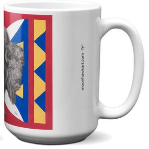 May include: White ceramic mug featuring a detailed illustration of a bison on a red, blue, and yellow geometric design. The mug has a large handle and the website address moonhawktart.com is printed on the side.