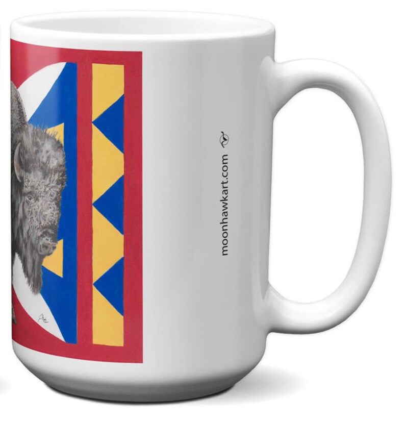 May include: White ceramic mug featuring a detailed illustration of a bison on a red, blue, and yellow geometric design. The mug has a large handle and the website address moonhawktart.com is printed on the side.