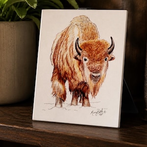May include: A square white tile featuring a detailed watercolor illustration of a brown bison standing in snow. The bison has dark curved horns and shaggy fur. The artist's signature, Mary Beth, is written in black ink at the bottom right corner.
