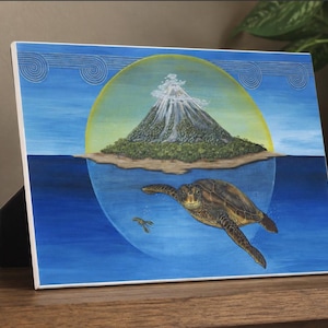 May include: A rectangular tabletop plaque featuring a painting of a sea turtle swimming in blue water below an island with a smoking volcano. The scene is framed by a yellow circular glow against a blue sky with stylized wave patterns at the top.