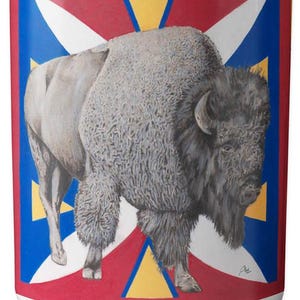 May include: White ceramic mug featuring a detailed illustration of a bison. The bison is depicted in shades of gray and brown against a backdrop of red, white, blue, and yellow geometric shapes. A unique and artistic design.