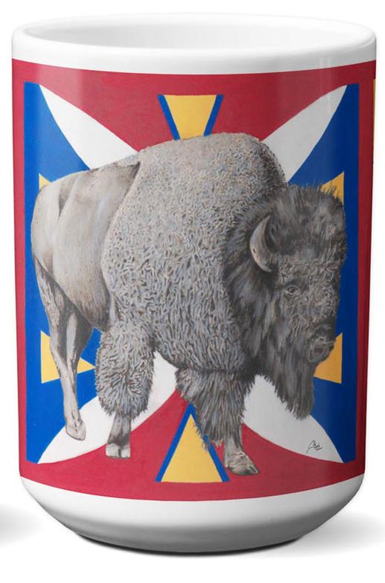 May include: White ceramic mug featuring a detailed illustration of a bison. The bison is depicted in shades of gray and brown against a backdrop of red, white, blue, and yellow geometric shapes. A unique and artistic design.