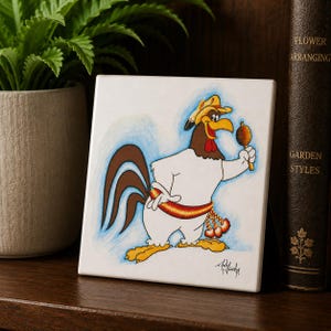 May include: A square ceramic tile featuring a cartoon illustration of a white rooster wearing a yellow hat and a red and orange patterned sash. The rooster holds a brown maraca in one wing. The artwork includes a signature in the bottom corner.