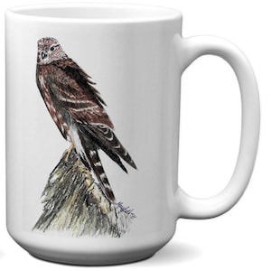 May include: White ceramic mug featuring a detailed illustration of a hawk perched on a rock. The hawk has brown and white feathers, and the rock is textured with shades of brown and gray. The mug has a large handle and a glossy finish.