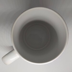May include: A white ceramic mug with a rounded interior and a handle on the side. The mug is empty and sits on a white surface. The mug's interior is a darker shade of gray.
