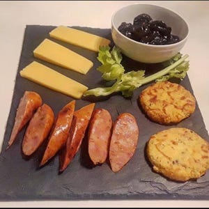 May include: A rectangular black slate serving board features three rectangular slices of yellow cheese, a white bowl filled with dark pitted olives, a sprig of green celery, six slices of cooked sausage, and two round golden-brown patties.