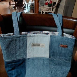 May include: A patchwork denim tote bag featuring various shades of blue fabric, two shoulder straps, and a light-colored interior lining with a small pattern. A rectangular patch reads Route 66 Original and a small brown leather tag is attached.