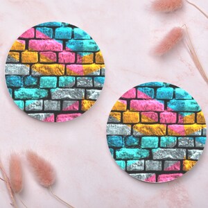 May include: Two round coasters with a brick wall design. The coasters are painted in shades of pink, blue, yellow, and gray.