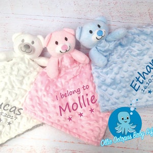 May include: Three personalized baby blankets with plush teddy bear attachments. The blankets are white, pink, and blue with embroidered names and dates. The pink blanket says "I belong to Mollie" and the blue blanket says "Ethan 16.10.2017".