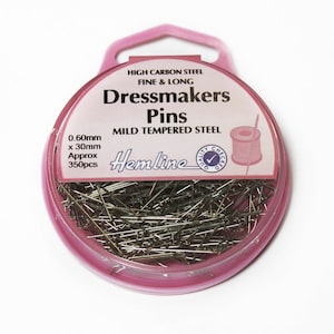 Könnte beinhalten: Ein rosa, kreisförmiger Behälter mit Schneiderstiften. Auf dem Etikett steht "Dressmakers Pins" und "Hemline". Die Stifte sind fein und lang, aus mildem, gehärtetem Stahl. Der Behälter gibt auch an, dass die Stifte 0,60 mm x 30 mm groß sind, mit ca. 350 Stück.