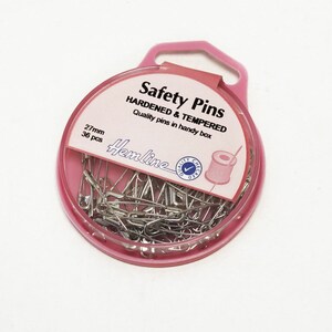 Könnte beinhalten: Ein rosa Plastikbehälter mit einem durchsichtigen Deckel, gefüllt mit 36 silbernen Sicherheitsnadeln. Der Behälter hat eine Aufhängeschlaufe und ein Etikett mit der Aufschrift "Safety Pins Hardened & Tempered Quality pins in handy box 27mm 36 pcs Hemline".