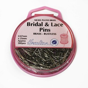 Könnte beinhalten: Ein rosa Plastikbehälter mit einem durchsichtigen Deckel, gefüllt mit 300 vernickelten Messing-Braut- und Spitzennadeln. Der Behälter hat eine Aufhängeschlaufe und ein Etikett mit der Aufschrift "Nickel Plated Brass Bridal & Lace Pins Brass - Rustless Hemline Quality Checked". Die Nadeln sind 0,67 mm x 25 mm groß.