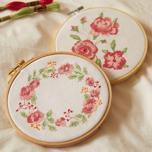Peut inclure: Deux cerceaux à broder avec des motifs floraux. Le cerceau de gauche présente une couronne de roses roses et de feuilles vertes. Le cerceau de droite présente un groupe de roses roses avec des feuilles vertes.