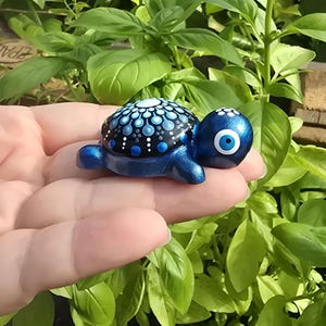 Könnte beinhalten: Eine kleine Schildkrötenfigur mit metallisch blauer Oberfläche und einem schwarzen Panzer, der mit einem weißen und hellblauen Punktmuster im Mandala-Stil verziert ist. Die Schildkröte hat ein blau-weißes Augensymbol an der Seite ihres Kopfes.