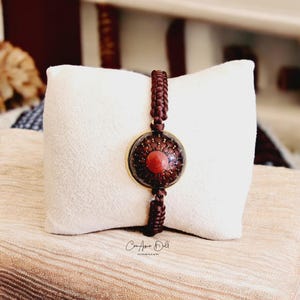 Può includere: Un braccialetto in corda marrone con un ciondolo rotondo con bordo dorato che presenta un design a mandala rosso e marrone.