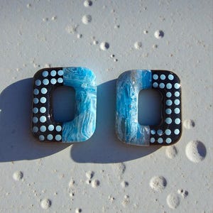 Può includere: Due orecchini quadrati. Ogni orecchino ha una sezione nera con pois azzurri e una sezione marmorizzata blu e bianca. Gli orecchini hanno un foro rettangolare al centro.