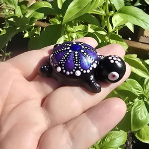 Könnte beinhalten: Eine kleine Schildkrötenfigur, schwarz bemalt mit einem leuchtend violetten und blauen Mandalamuster auf dem Panzer. Der Panzer weist detaillierte weiße Punktverzierungen auf. Die Schildkröte hat einen schwarzen Kopf mit einem rosa Auge.