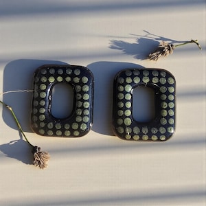 Può includere: Due orecchini rettangolari neri con apertura centrale, decorati con un motivo a piccoli pois verde chiaro. Gli orecchini sono esposti su una superficie chiara, con uno stelo di fiore secco nelle vicinanze.