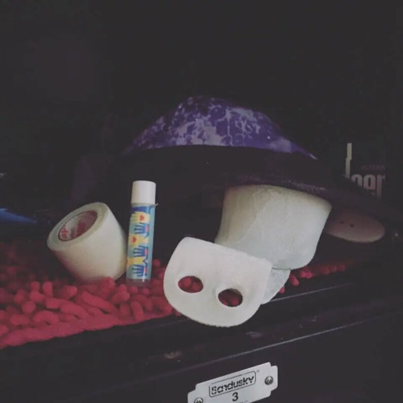 May include: A white athletic tape roll, a small blue and white tube of athletic tape, and a white athletic wrist brace with two holes in it. The items are in a black drawer with a red fuzzy mat in the background. The drawer has a metal label that says "Sandusky 3".